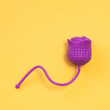 Infusor de té de silicona en forma de rosa color morado sobre fondo amarillo.